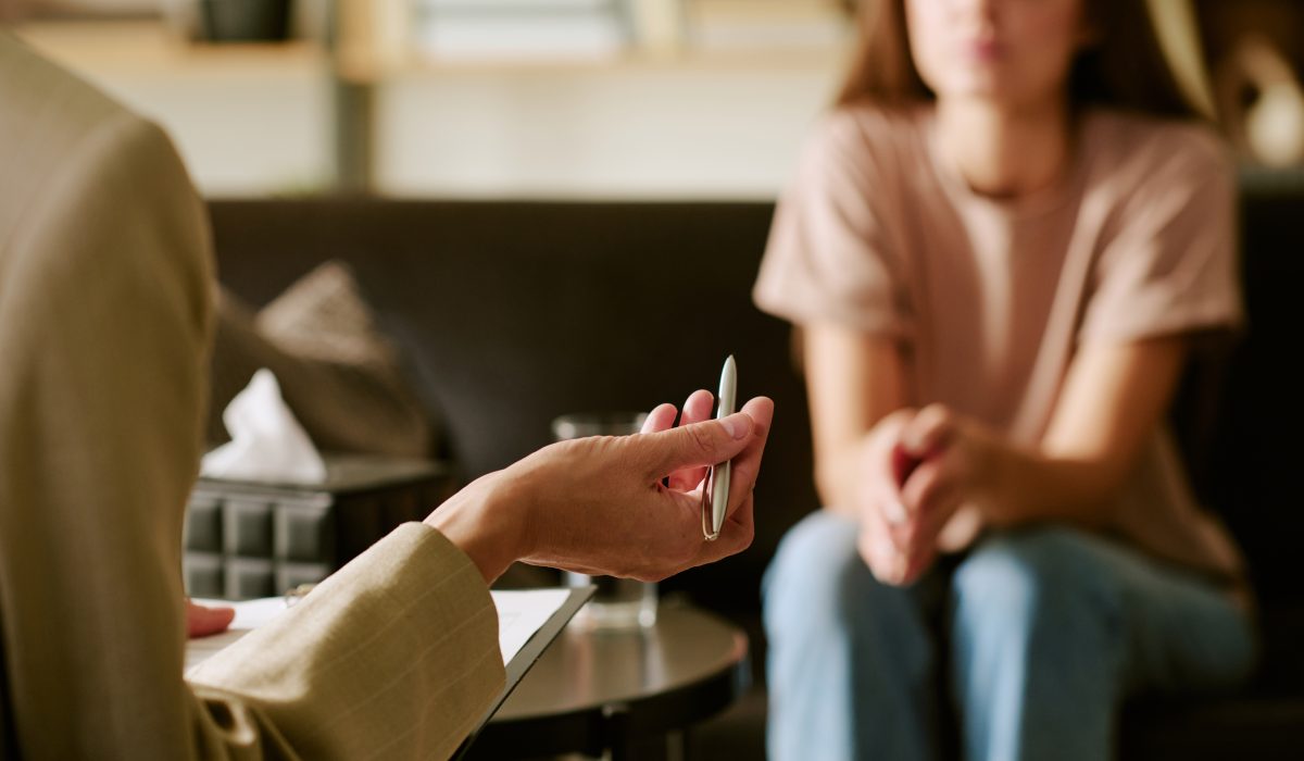 Caucasian young adult woman sitting on couch listening to middle aged Caucasian man holding pen during psychological counseling session in office setting
