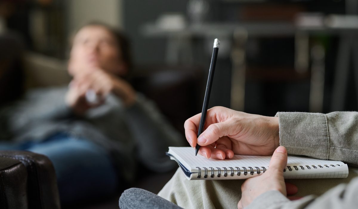 Therapist holding pen and notepad while patient sits in background with blurred face. Focus is on therapist's hand and pen without showing patient's details